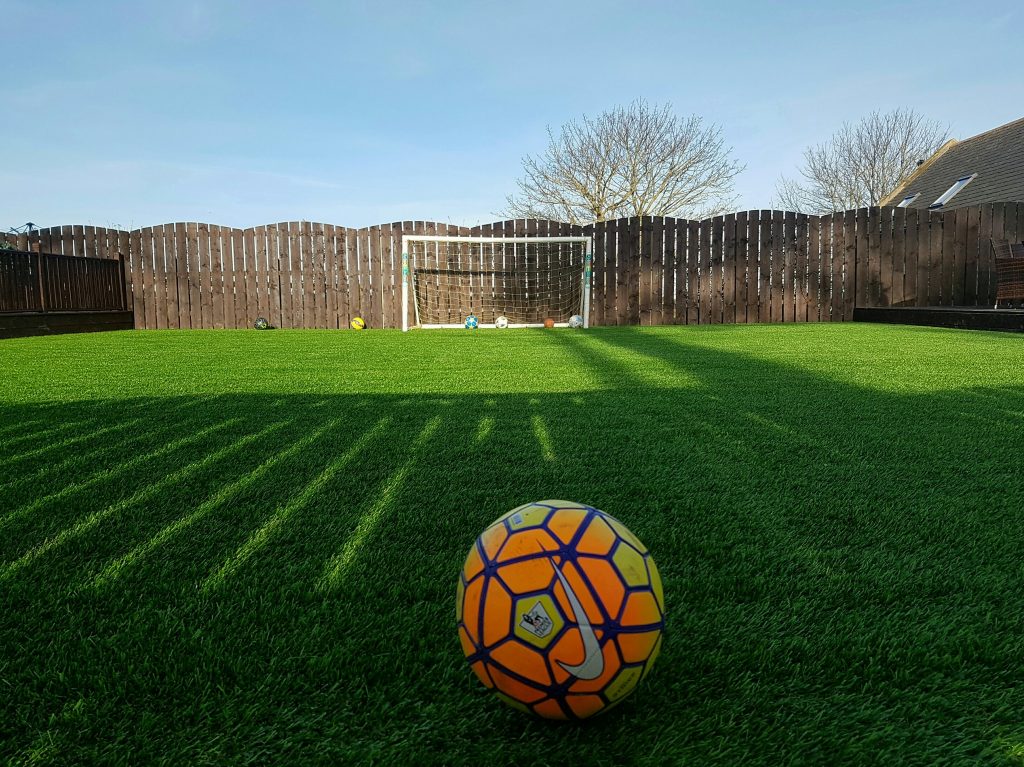 A football in the foreground on artificial grass with a small football goal and 6 other footballs in the background