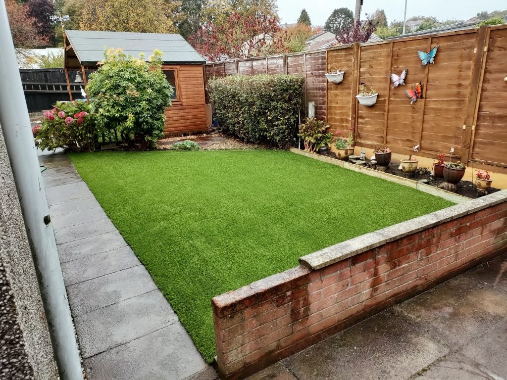 A garden lawn fitted with artificial grass, surrounded by flower pots, hedges and a wooden shed at the bottom of the garden